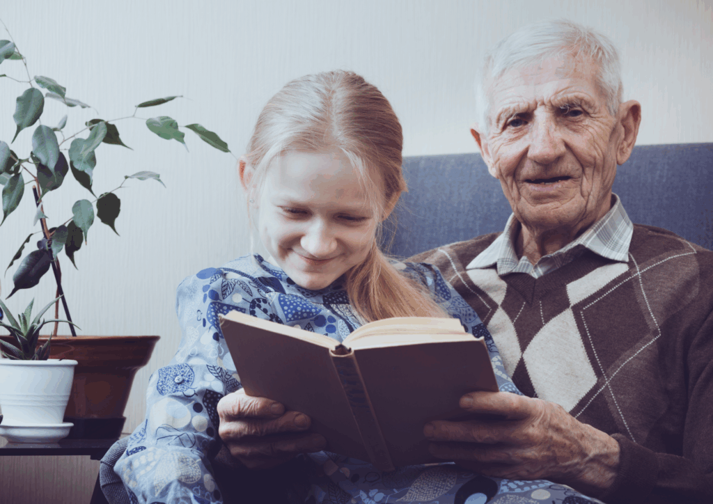 An older grandfather reading a book with his granddaughter. Both are smiling.