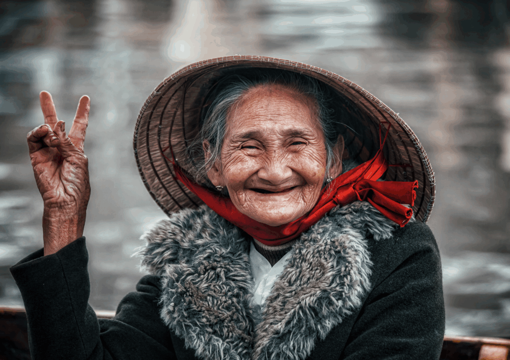 An older woman throwing up a peace sign and smiling while wearing a sun hat with a red scarf.