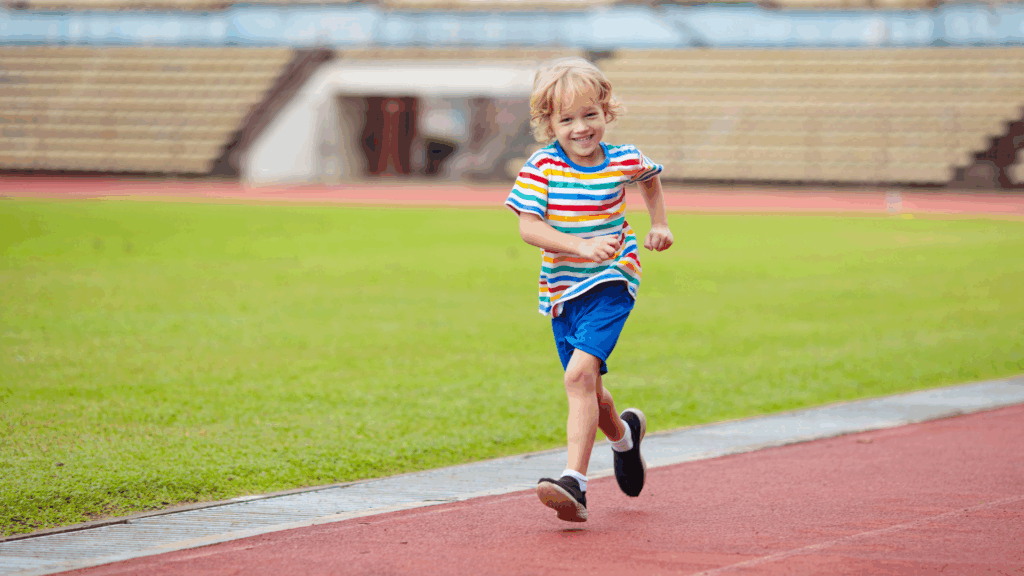 A blonde kid with a striped shirt and blue shorts running on a track.